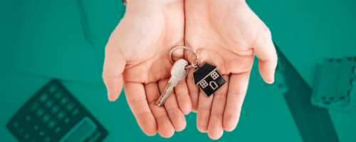 The picture shows hands holding keys with a keychain in the shape of a house. In the background, a calculator and documents are visible.