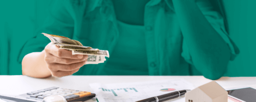A woman sits at a table, holding money in her hand. On the table lie documents, credit cards, a calculator, a small car and a house stand.