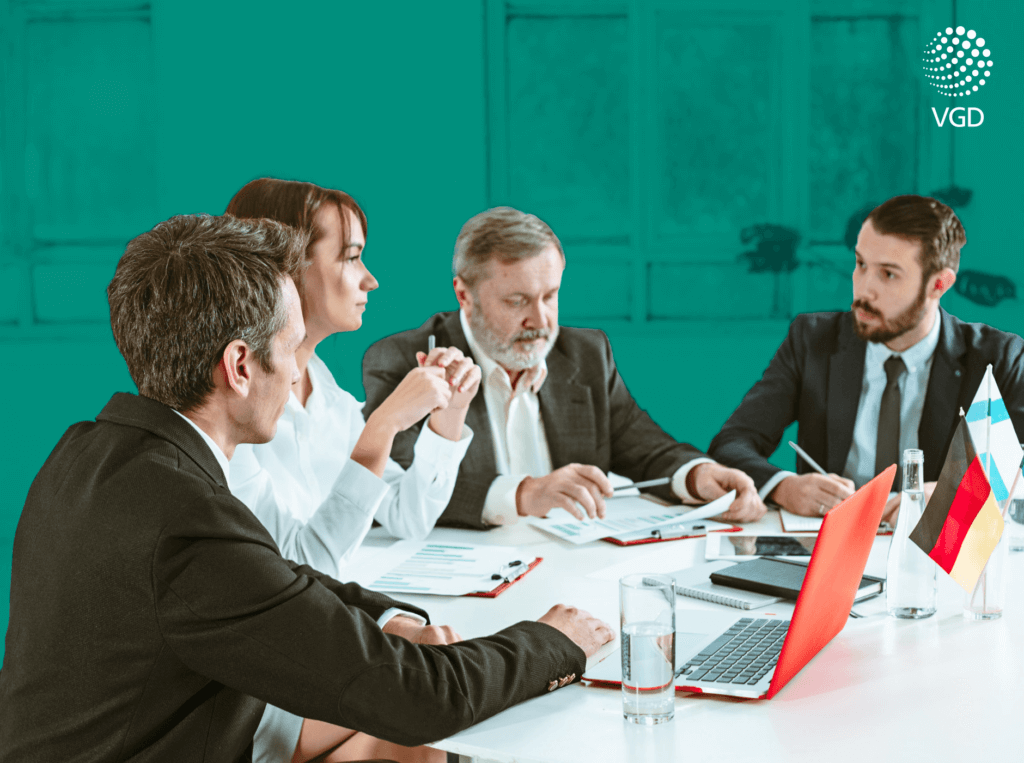 A four-person business group discussing documents at a conference table, with a laptop and the flags of Germany and Finland.