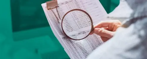 Osoba przeglądająca śródroczne sprawozdanie finansowe przy użyciu lupy, w tle biurko z komputerem.