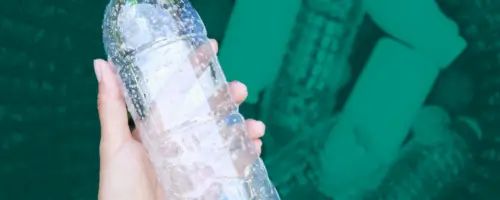 na zdjęciu jest ręka trzymająca butelkę plastikową, w tle jest kosz z większą ilością plastikowych butelek.