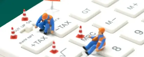Miniaturowi drogowcy w kamizelkach pracują na klawiaturze komputerowej, gdzie widoczne są klawisze oznaczone TAX+ i TAX-, symbolizujące zmiany w podatkach.